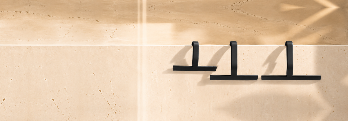 Three matte black dancemoon JustHang shower squeegees—Compact, standard and Wider—hanging on the right side of a sunlit beige stone wall.