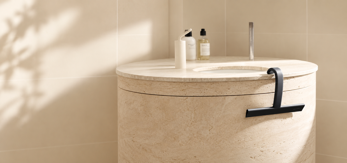 Black dancemoon JustHang shower squeegee with built-in hook on a beige stone bathroom vanity.